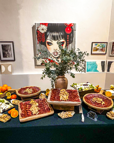 Mesa de ibéricos gourmet | Catering en Valencia Mesa decorada con platos de comida, frutas y una obra de arte en la pared.